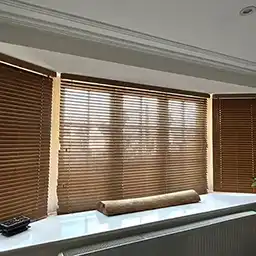 Wooden blinds cover large windows in a bright room, with sunlight filtering through. A smooth, light-colored surface is in front.